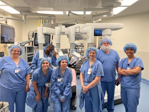 A group of Surgical Technician students wearing blue scrubs and hair covers pose together in an operating room setting, surrounded by surgical equipment and robotic technology.