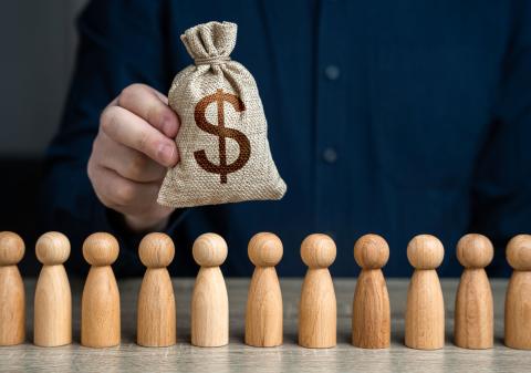 A person holds a small burlap bag with a dollar sign above a row of wooden figures, symbolizing investment in people or workforce development.